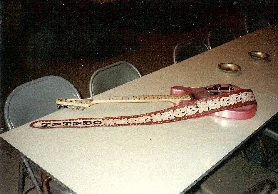 A pink paisley Fender Telecaster lying on a folding table with its tooled leather strap coiled beside it.