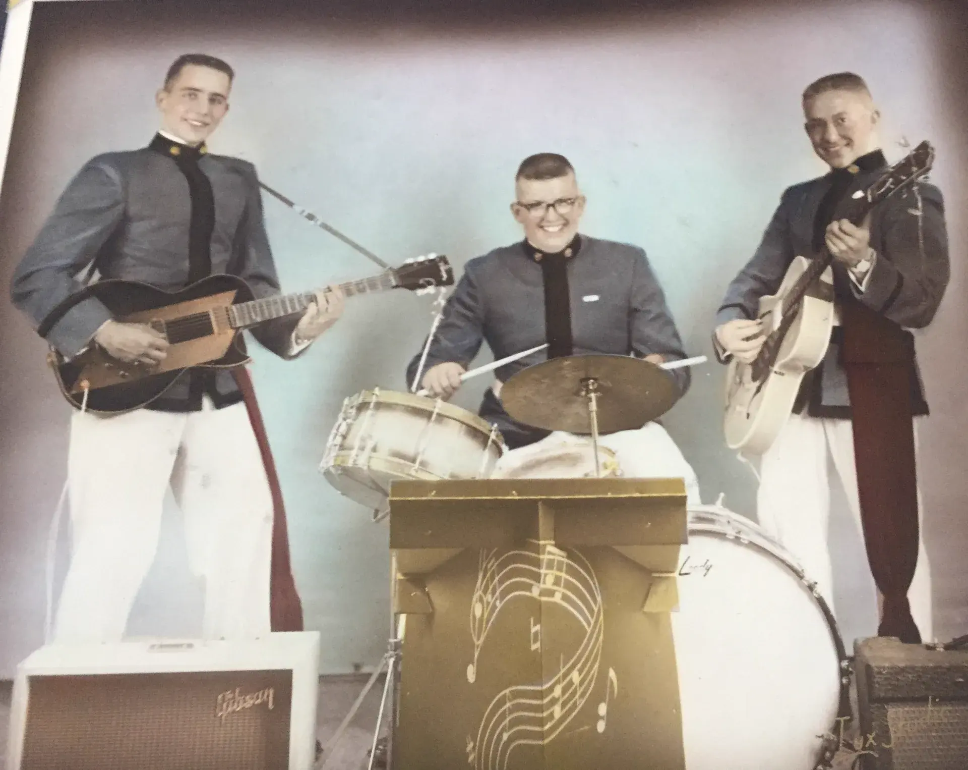 Hand-coloured studio portrait of three young men in grey military academy uniforms and white trousers with two archtop guitars and a drum kit, behind a gold bandstand painted with a treble clef.