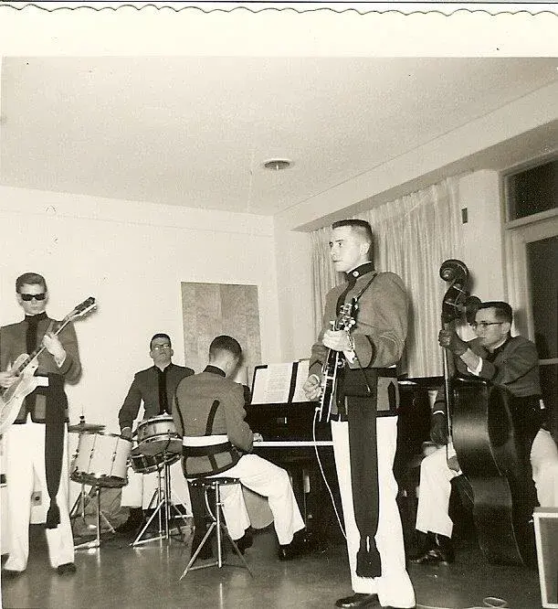 Black and white photograph of six young men in military academy uniforms playing a function room: two guitars, drums, piano and upright bass.