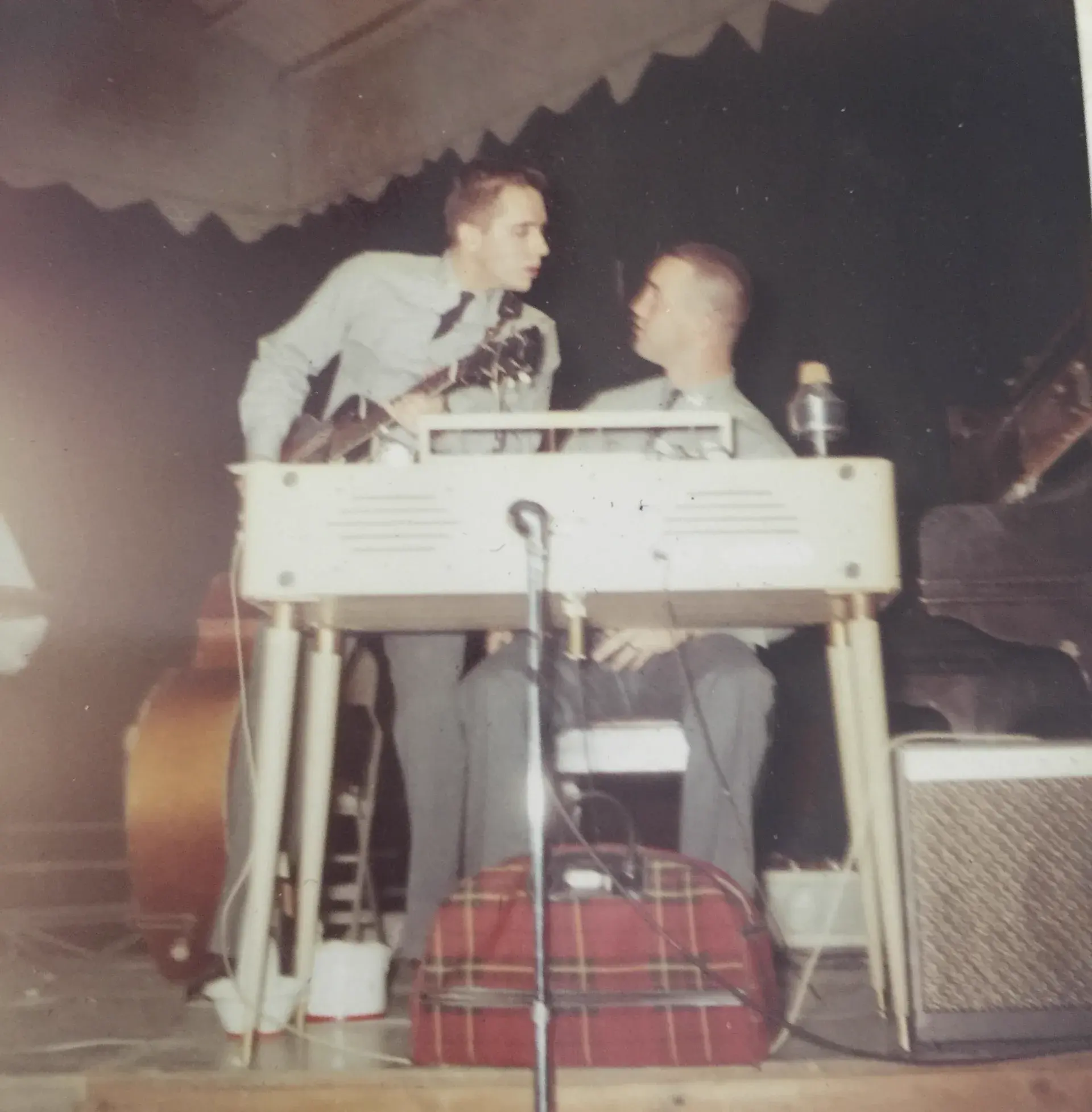 Two young men in military academy uniforms under a marquee, one leaning over a white pedal steel guitar, a tartan bag and amplifier at their feet.