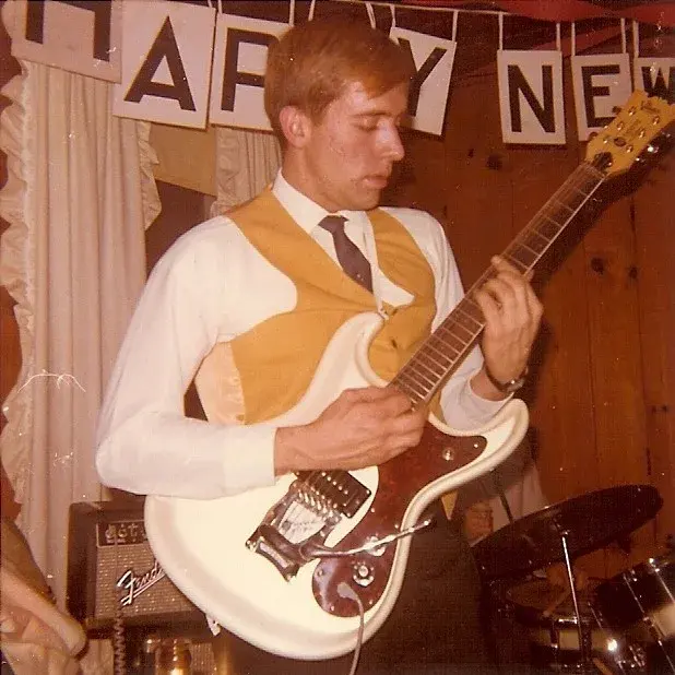 A young man in a mustard waistcoat and tie playing a white offset-body Fender guitar beneath a New Year banner, a Fender amplifier behind him.