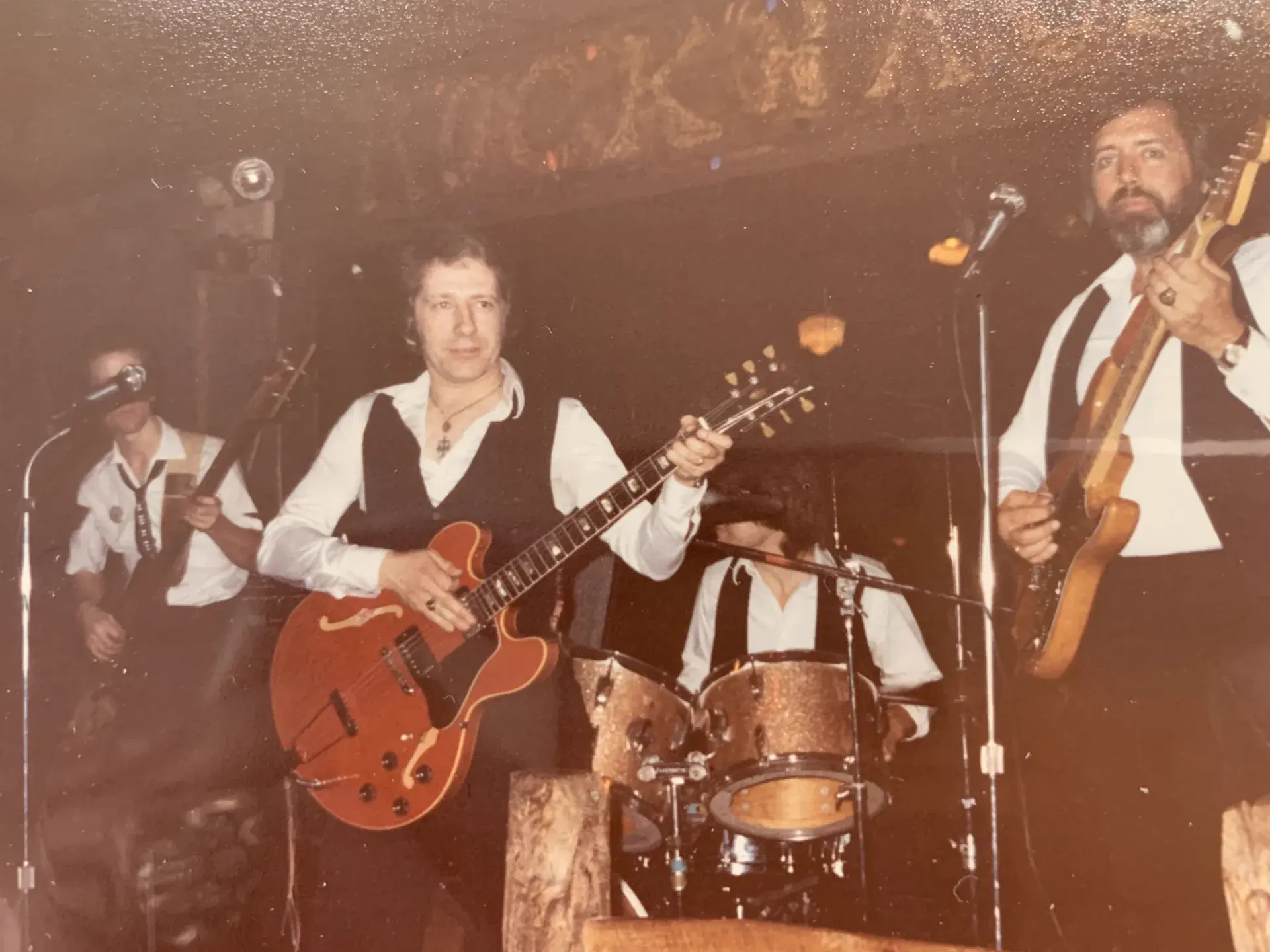 A band in white shirts, black waistcoats and ties performing beneath a "Rock On" sign, the guitarist at centre playing a red Gibson ES-335, a glitter drum kit behind.