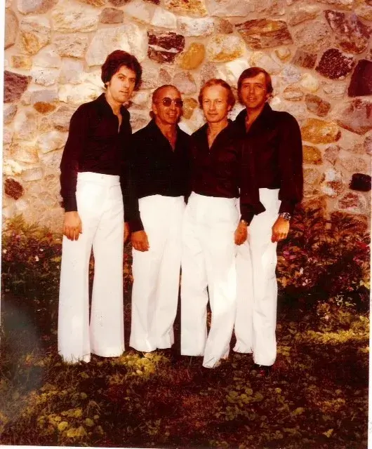 Four men in black shirts and white flared trousers posed against a fieldstone wall.