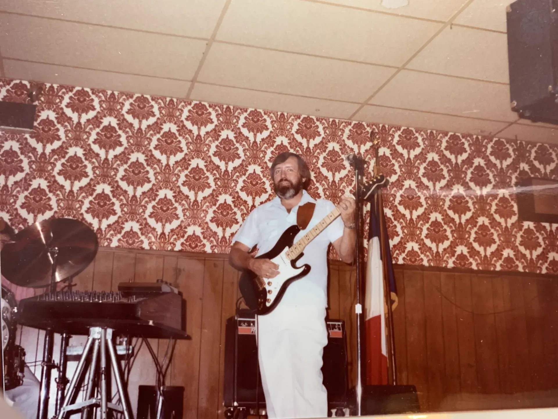A bearded guitarist dressed in white playing a black Stratocaster on a small stage with red damask wallpaper, a pedal steel guitar to his left and a French tricolour flag on a stand.