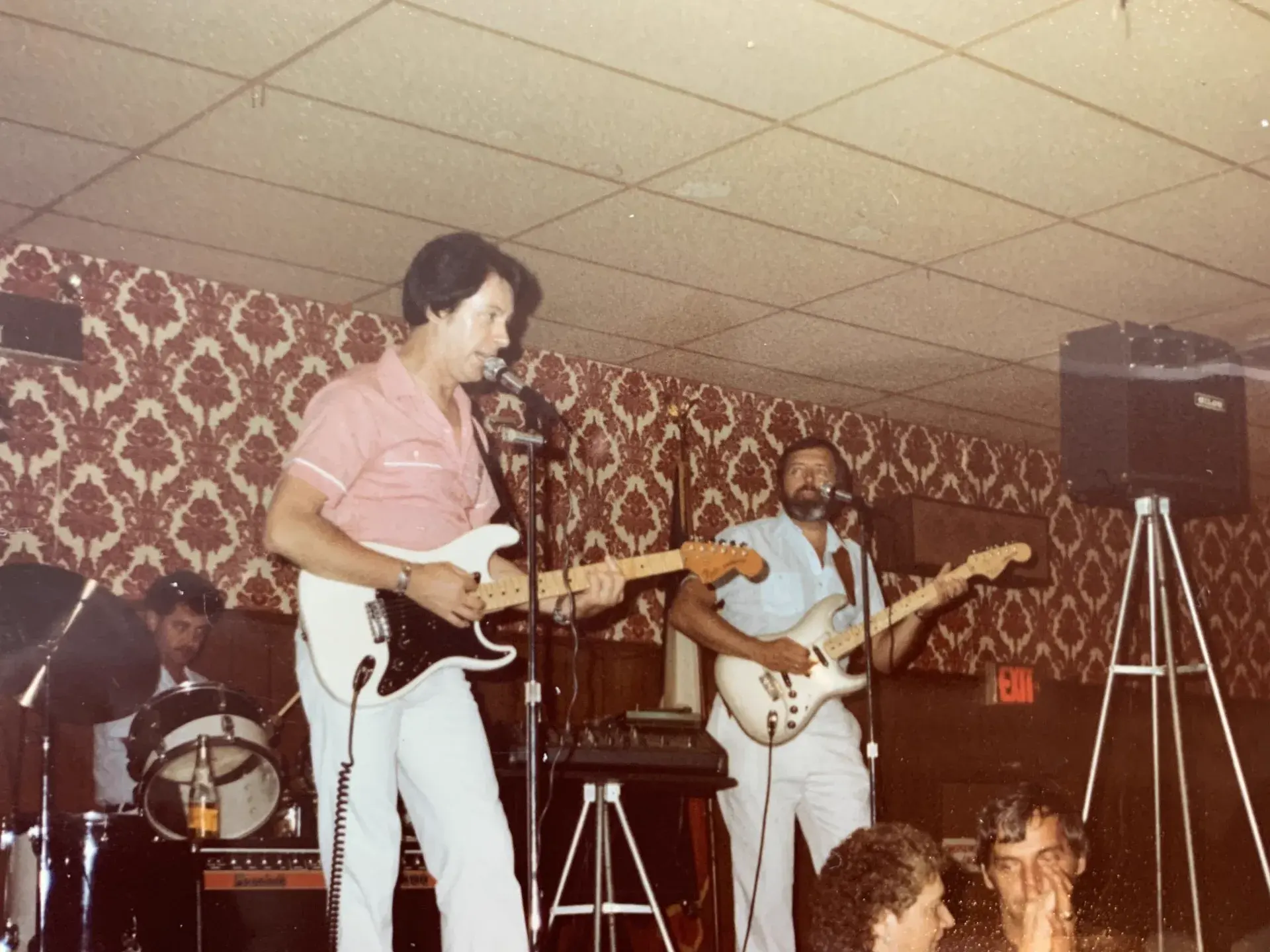 Two guitarists on a damask-papered stage, one singing in a pink shirt with a black-guard Stratocaster, the other on a white Stratocaster, drummer behind and audience in the foreground.