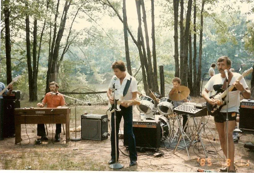 A country band playing outdoors among trees beside water in August 1985: pedal steel, Yamaha keyboard, drums and two guitars.