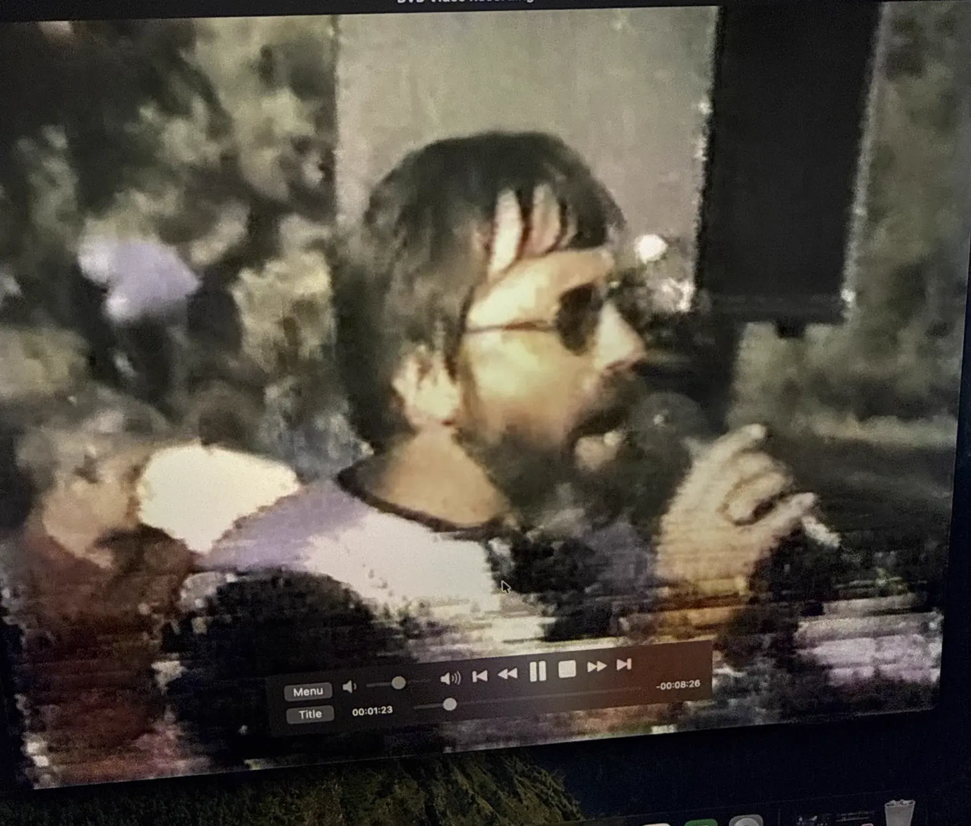 A photograph of a screen showing archival video: a bearded man in sunglasses at an outdoor event, DVD player controls visible along the bottom of the frame.