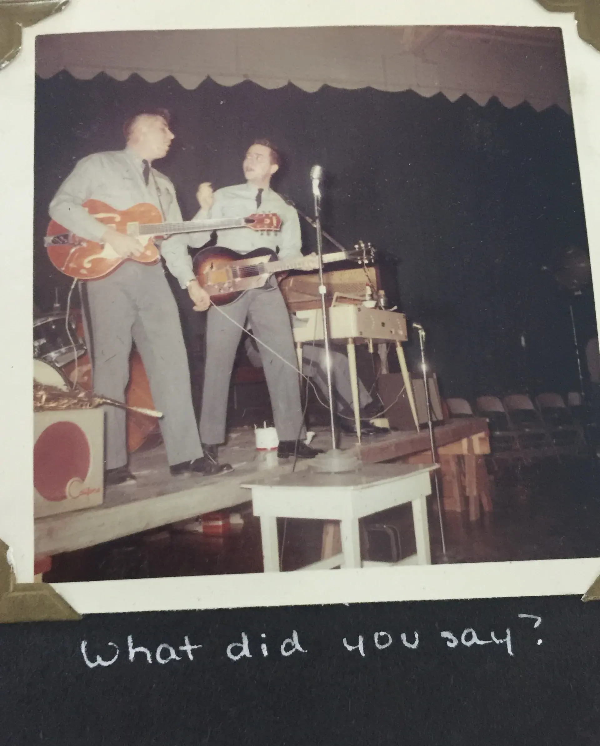 Album page: two guitarists in matching grey shirts and dark ties on a low stage, one with an orange archtop, handwritten caption reading What did you say?