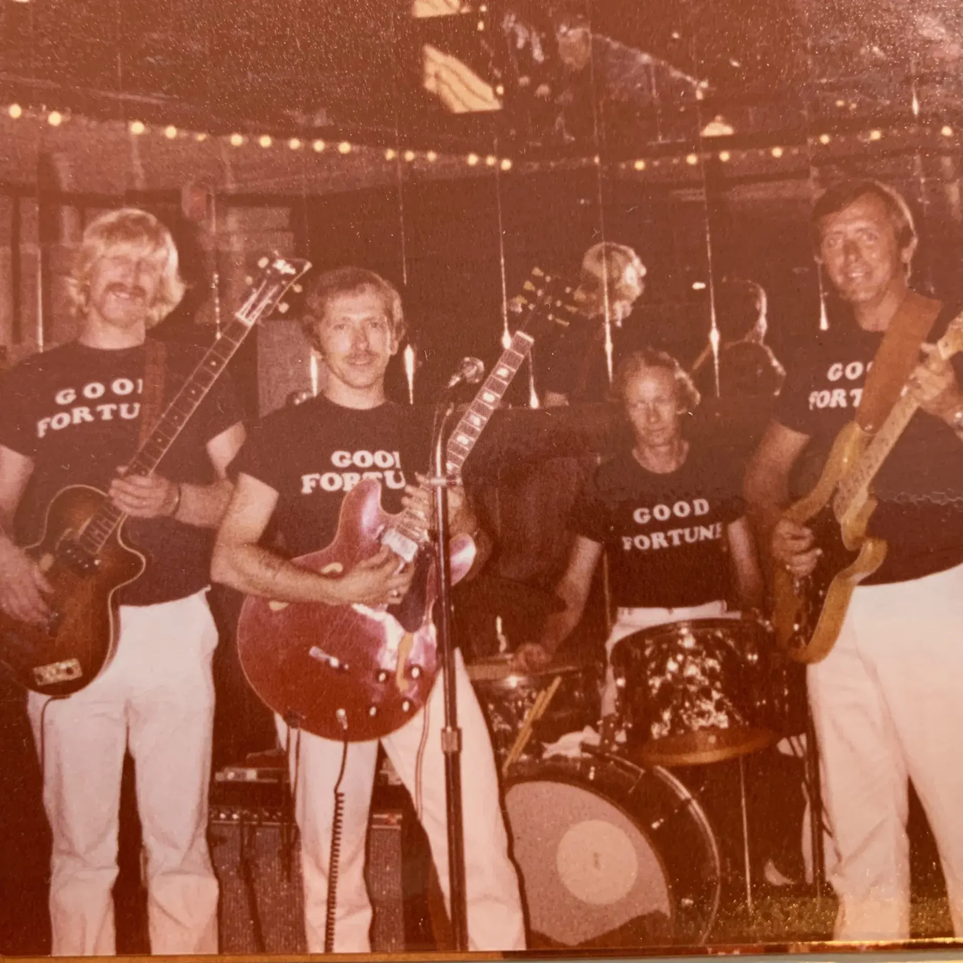 Five-piece band on stage in matching GOOD FORTUNE t-shirts and white trousers under festoon lights, one player holding a cherry-red Gibson ES-335.