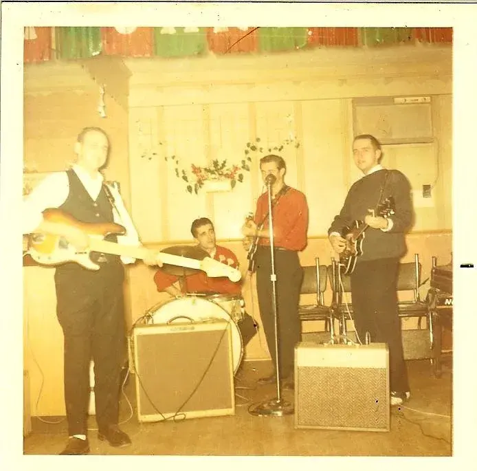 A four-piece band playing a small hall dressed with holiday streamers: blond Fender bass, drums, a singer in a red shirt, and a guitarist with a dark solidbody.