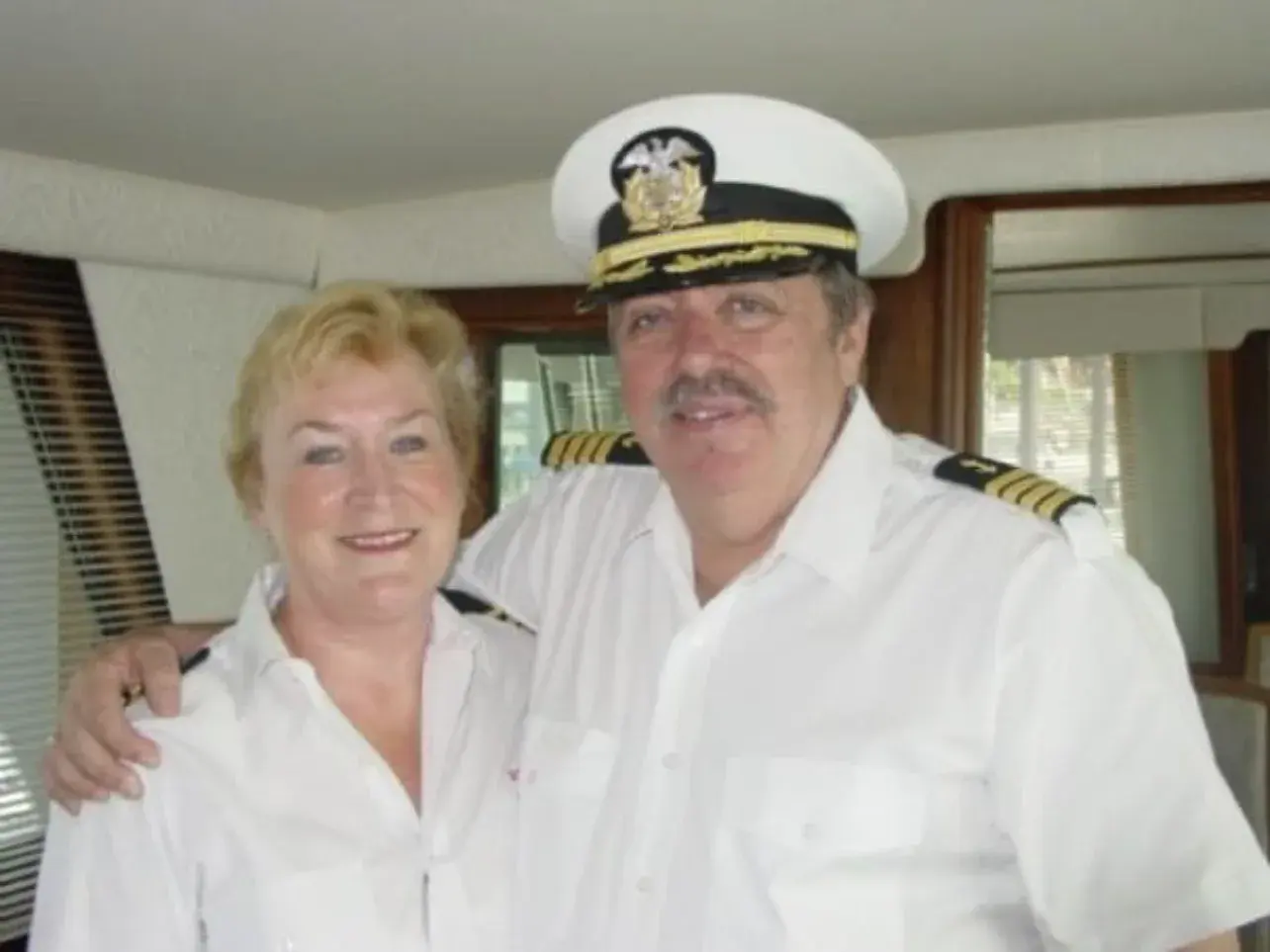 Graham and Bonnie Nicholson in the wheelhouse, both in white crew shirts with epaulettes, Graham in a captain's peaked cap.