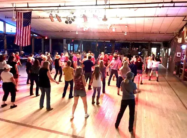 Couples line dancing on the Cadillac Ranch floor beneath American flags and stage lighting.
