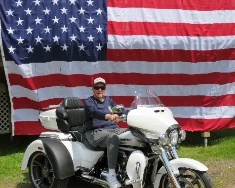 A rider on a white Harley-Davidson trike in front of a large American flag.