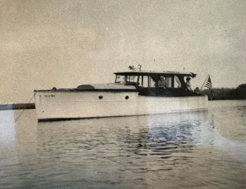 A black-and-white photograph of a wooden cabin cruiser underway on calm water.