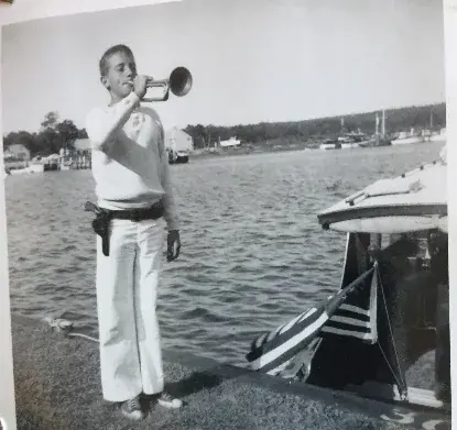 A man in whites aboard a small boat raising a bugle to his lips, moored boats behind him.