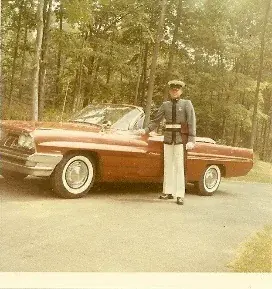 A young man in military-academy uniform standing beside a red convertible on a wooded drive.