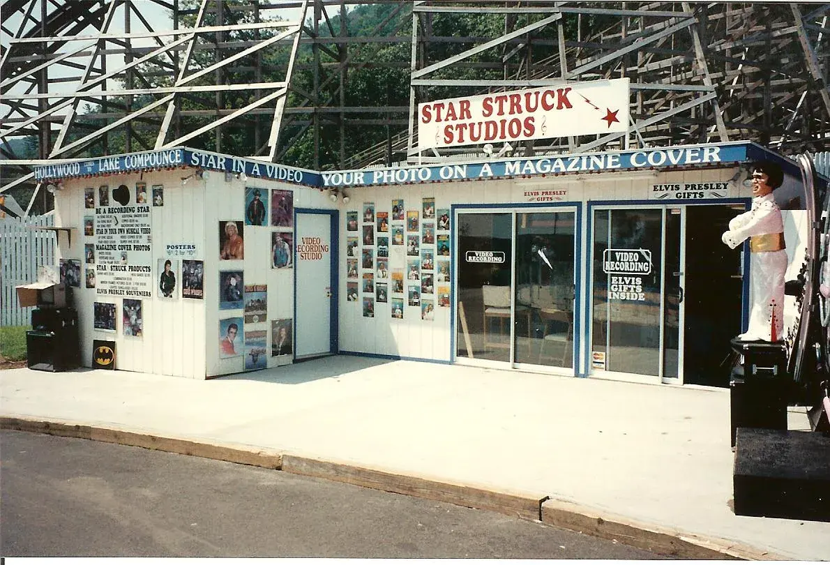 Star Struck Studios at Lake Compounce — Graham Nicholson's video and karaoke booth, a white building beneath a wooden roller coaster, with signs reading "Star in a video, your photo on a magazine cover" and "Elvis Presley Gifts" and an Elvis statue by the door.